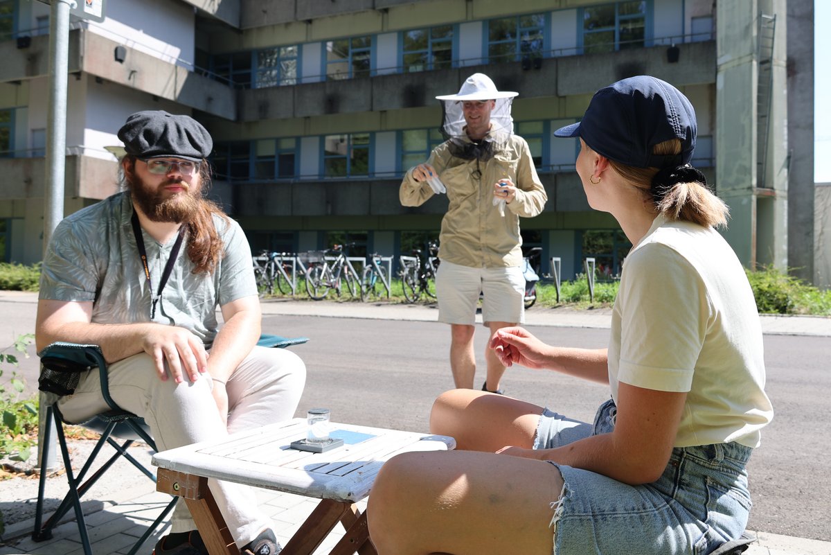 Eine Person trägt einen Imker-Helm. Zwei Personen warten sitzend auf ihn und die Bienen, die er mitbringt.